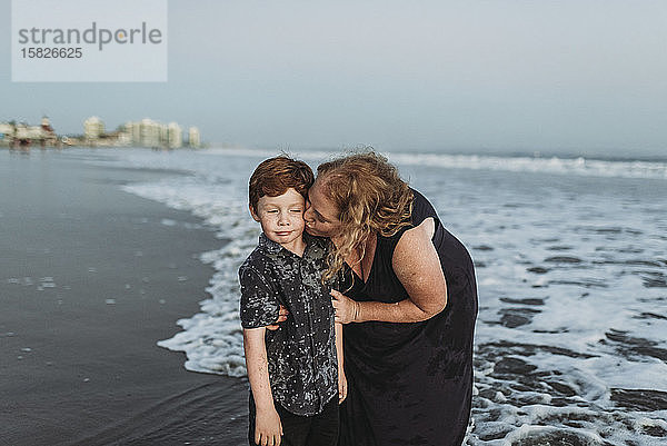 Sohn macht ein lustiges Gesicht  während Mutter ihn in der Abenddämmerung am Strand küsst