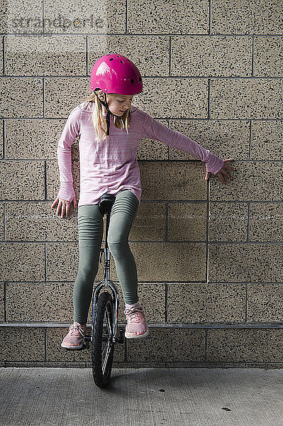 Mädchen mit rosa Helm balanciert auf einem Einrad gegen eine Ziegelmauer