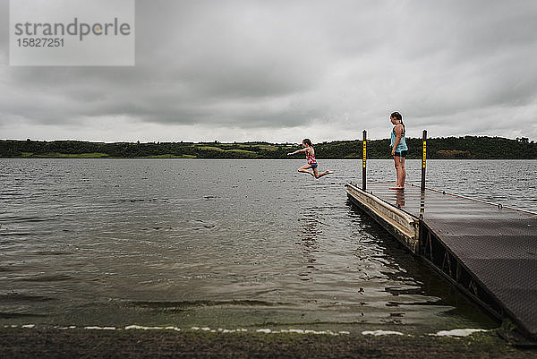 Kleine Mädchen springen an einem bewölkten Sommertag vom Dock in den See