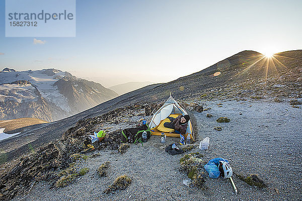 Mann stößt bei Sonnenuntergang den Kopf aus dem Zelt  während er auf einem Bergrücken zeltet.