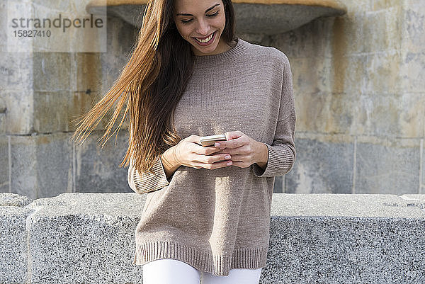 Lächelnde Frau benutzt ihr Telefon  während sie sich an einen Brunnen in Boadilla lehnt