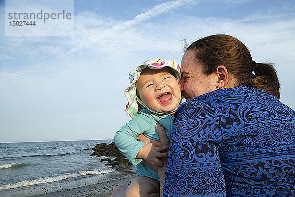 Mutter umarmt glücklich lächelndes Mädchen am Strand sitzend