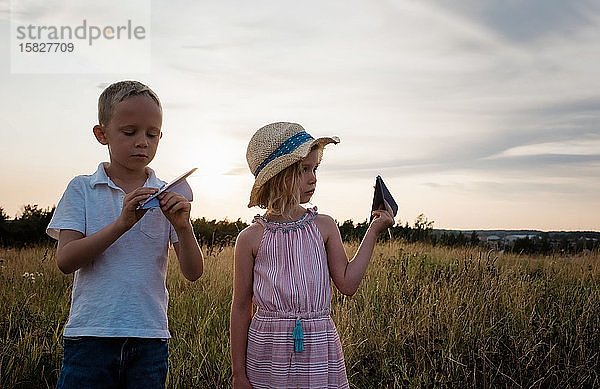 Geschwister spielen mit Papierflugzeugen auf einer Wiese bei Sonnenuntergang