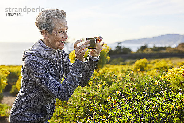 Fröhliche ältere Frau fotografiert mit einem Smartphone  während sie bei Pflanzen gegen den Himmel steht