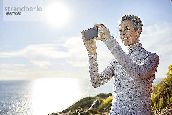 Lächelnde ältere Frau fotografiert durch ein Smartphone  während sie an einem sonnigen Tag am Himmel steht