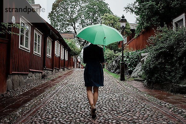 der Rücken einer Frau  die mit einem Regenschirm eine gepflasterte Straße hinaufgeht