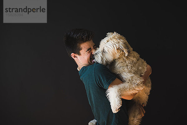 Teenager-Junge  der einen flauschigen Hund hält  der ihm das Gesicht ableckt.