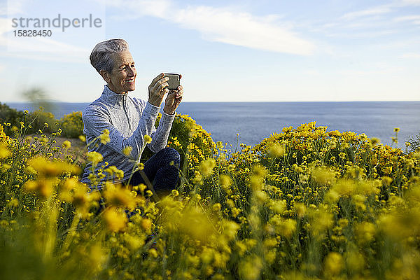 Lächelnde ältere Frau fotografiert mit einem Smartphone  während sie am Meer bei Pflanzen gegen den Himmel kauert