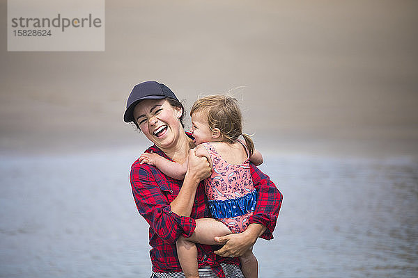 Mutter lacht  während sie ein kleines Mädchen am Strand hält.