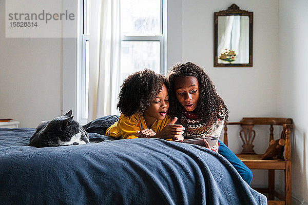 Mädchen zeigt Schwester Smartphone  während sie zu Hause von Katze auf dem Bett liegt