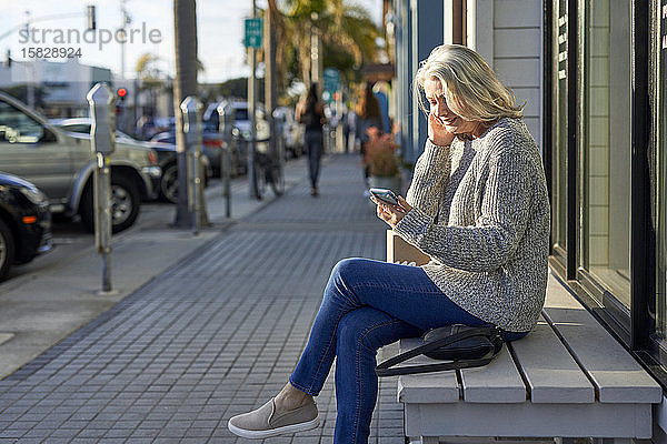 Seitenansicht einer älteren Frau  die ein Smartphone benutzt  während sie in der Stadt auf einer Bank sitzt