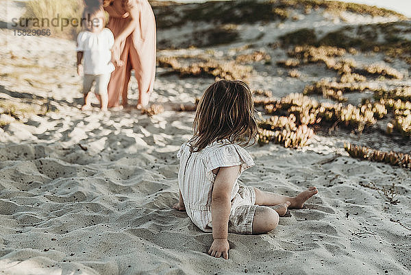 Hinteransicht eines jungen Kleinkindes  das im Sand sitzt und die Familie betrachtet
