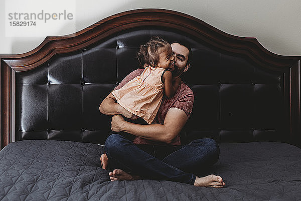Barfüßiger Papa sitzt im Bett und lacht mit seiner 2 Jahre alten Tochter