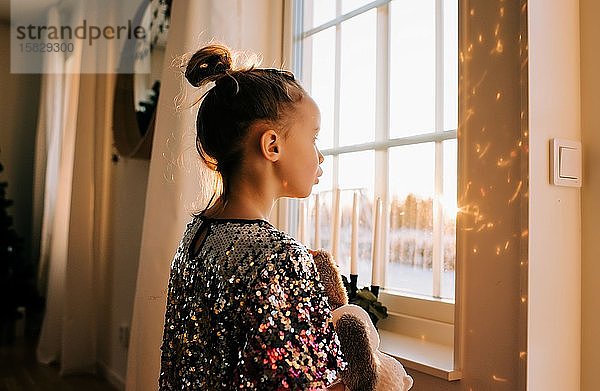 Junges Mädchen spielt mit ihrem Spielzeug zu Hause in einem glitzernden Kleid bei Sonnenuntergang