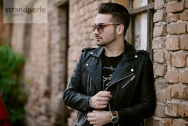 Trendiger Mann mit Sonnenbrille in einer Jacke  der an einer Backsteinmauer steht.