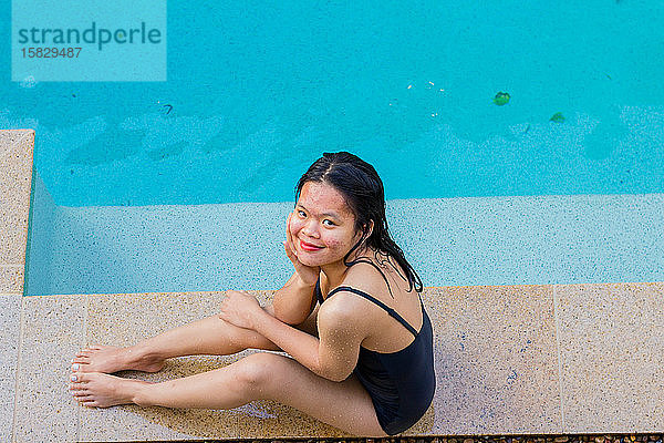 Schöne asiatische Frau sitzt am Poolrand an einem sonnigen Tag und lächelt