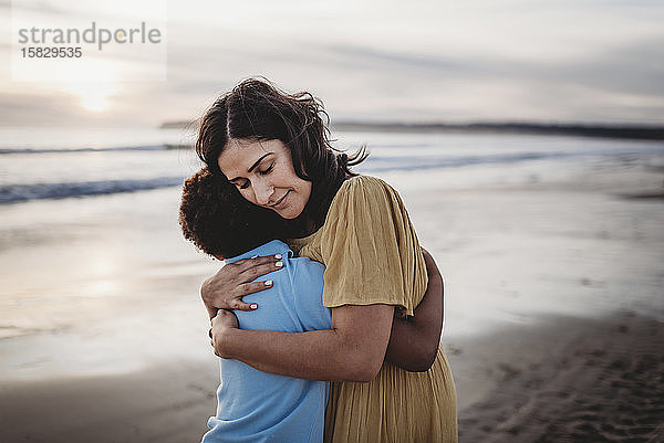 Seitenansicht einer schönen Mutter  die ihren Sohn bei Sonnenuntergang am Meeresufer umarmt