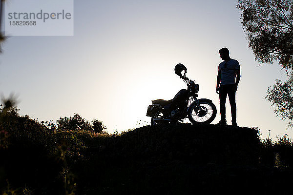 Hintergrundbeleuchtung eines Mannes  der sein schönes Motorrad auf einem Felsen in der