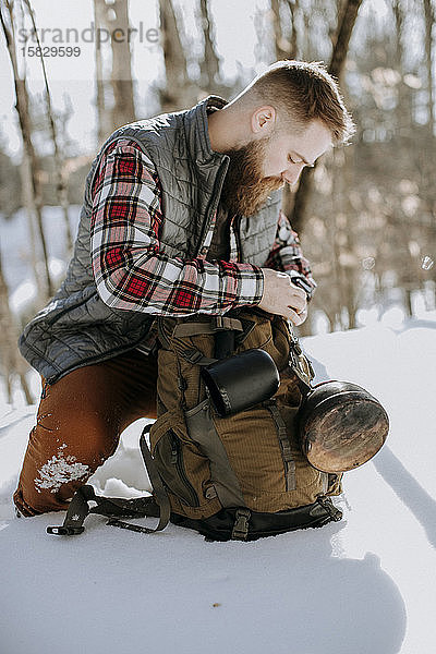 Mann mit Wulst in Flanell und Weste kniet mit Rucksack im Schnee