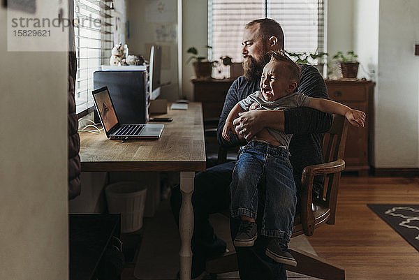 Vater arbeitet von zu Hause aus mit einem einjährigen Jungen  der auf dem Schoß weint