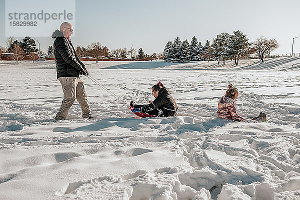 Seitenansicht eines Mannes  der im Winter zwei Mädchen auf einem Schlitten durchs Feld zieht