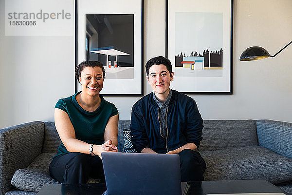 Porträt von lächelnden Geschäftskollegen  die mit Laptop am Tisch an der Wand im Büro sitzen