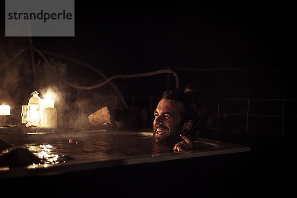 Mann mit Bart lacht in dampfender Außenbadewanne mit Kerzen