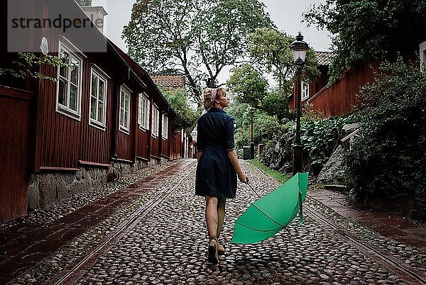 Frau  die auf einer gepflasterten Straße mit einem Regenschirm geht und den Regen betrachtet