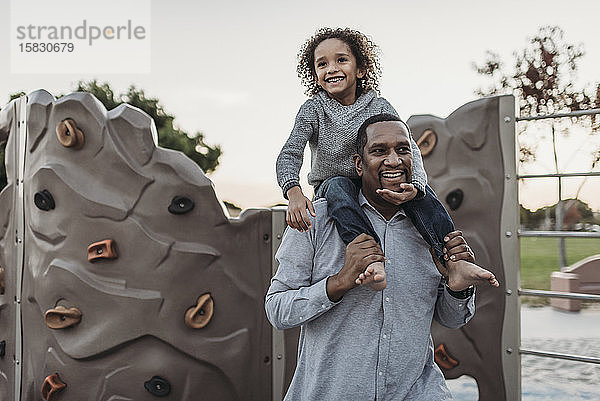 Fröhlicher Sohn sitzt auf den Schultern eines glücklichen Vaters auf einem Spielplatz im Park