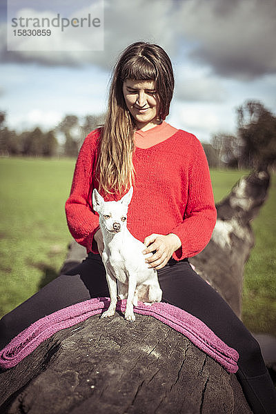 Frau in knallrotem Pullover sitzt mit kleinem weißen Hund auf einem Baumstamm auf dem Bauernhof