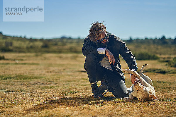 Porträt eines jungen tätowierten Mannes  der mit seinem Hund auf dem Land spielt