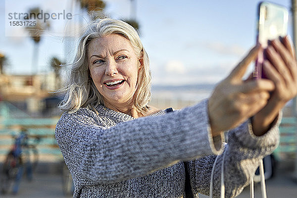 Lächelnde ältere Frau  die sich bei Sonnenuntergang am Strand mit einem Smartphone selbstständig macht