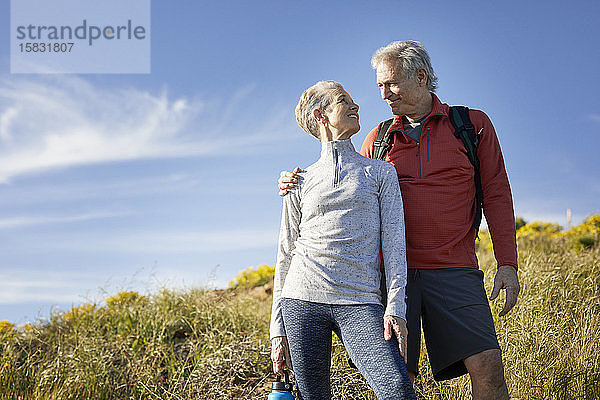 Niedrigwinkelansicht eines lächelnden älteren Paares  das sich gegenseitig ansieht  während es an einem sonnigen Tag auf einer Klippe vor blauem Himmel steht