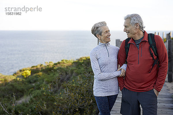 Lächelndes älteres Ehepaar  das sich Arm in Arm anschaut  während es auf einer Strandpromenade am Meer gegen den Himmel steht