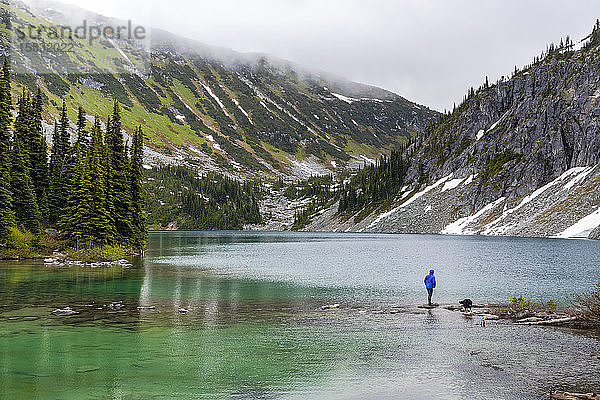 Eine Frau hängt mit ihrem Hund am Ufer eines Alpensees herum und nimmt an einem verregneten Sommertag die Aussicht in den Coast Mountains um Pemberton  British Columbia  auf.