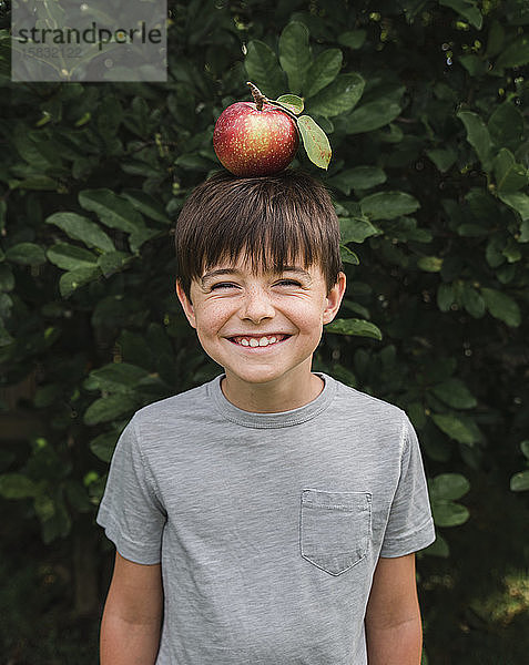 Glücklicher Junge  der draußen an einem Baum einen Apfel auf dem Kopf balanciert.