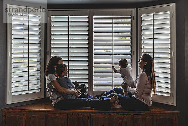 Gleichgeschlechtliche Eltern faulenzen mit Kindern auf einem Fensterplatz mit hohen Fensterläden