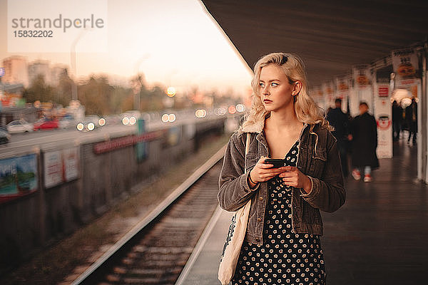 Junge nachdenkliche Frau benutzt Smartphone beim Warten auf den Zug