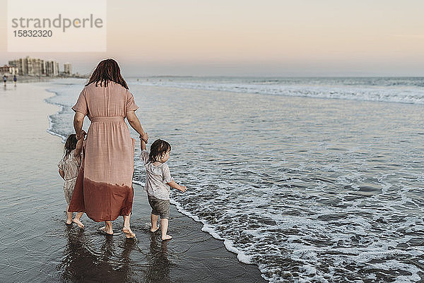 Hinteransicht einer Mutter  die junge Töchter im Meer am Strand spazieren führt