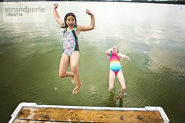 Zwei junge Mädchen in Badeanzügen springen von einem Dock in einen See