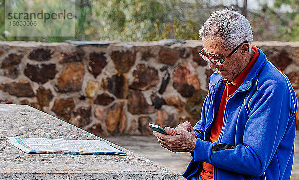 Älterer Mann schaut und berührt Bildschirm eines Smartphones  während er im Park sitzt