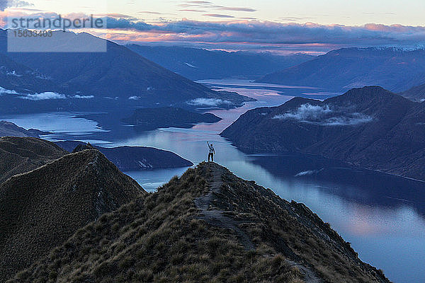 Mann steht bei Sonnenuntergang triumphierend auf einem Berggipfel in Neuseeland.