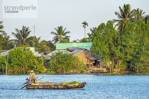Eine Frau in einem Boot passiert ein Dorf an einem Seitenarm des Mekong