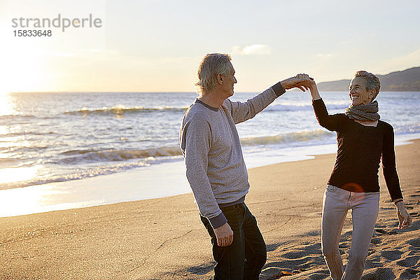 Lächelndes älteres Paar tanzt bei Sonnenuntergang am Strand