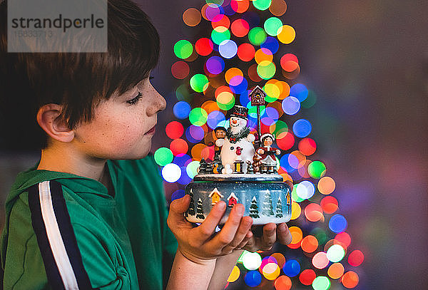 Junge hält Weihnachtsschmuck mit Baumlichtern im Hintergrund.