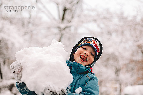 Glücklicher Junge streckt beim Spielen mit Schnee im Winter die Zunge heraus