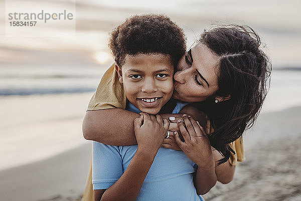 Nahaufnahme einer Mutter  die einen Jungen im Schulalter bei Sonnenuntergang am Strand küsst