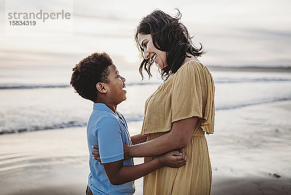 Seitenansicht einer schönen Mutter  die ihren Sohn bei Sonnenuntergang am Meeresufer umarmt
