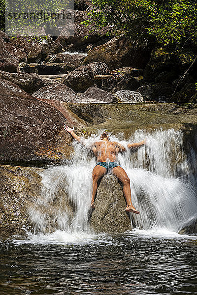 Junger erwachsener Mann sitzt über einem Felsen und badet am Regenwald-Wasserfall