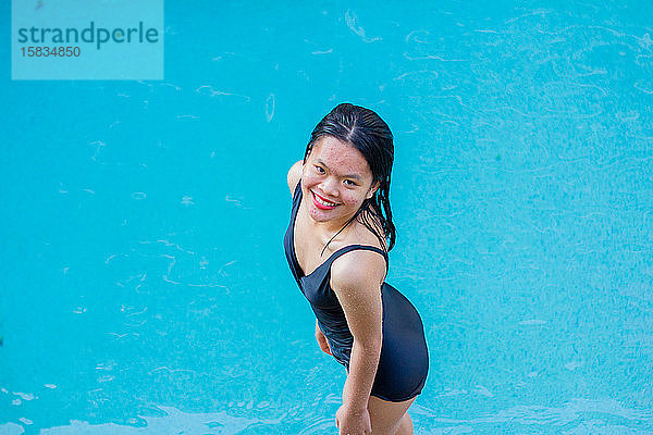 Schöne asiatische Frau lächelt im Stehen am Poolrand an einem sonnigen Tag